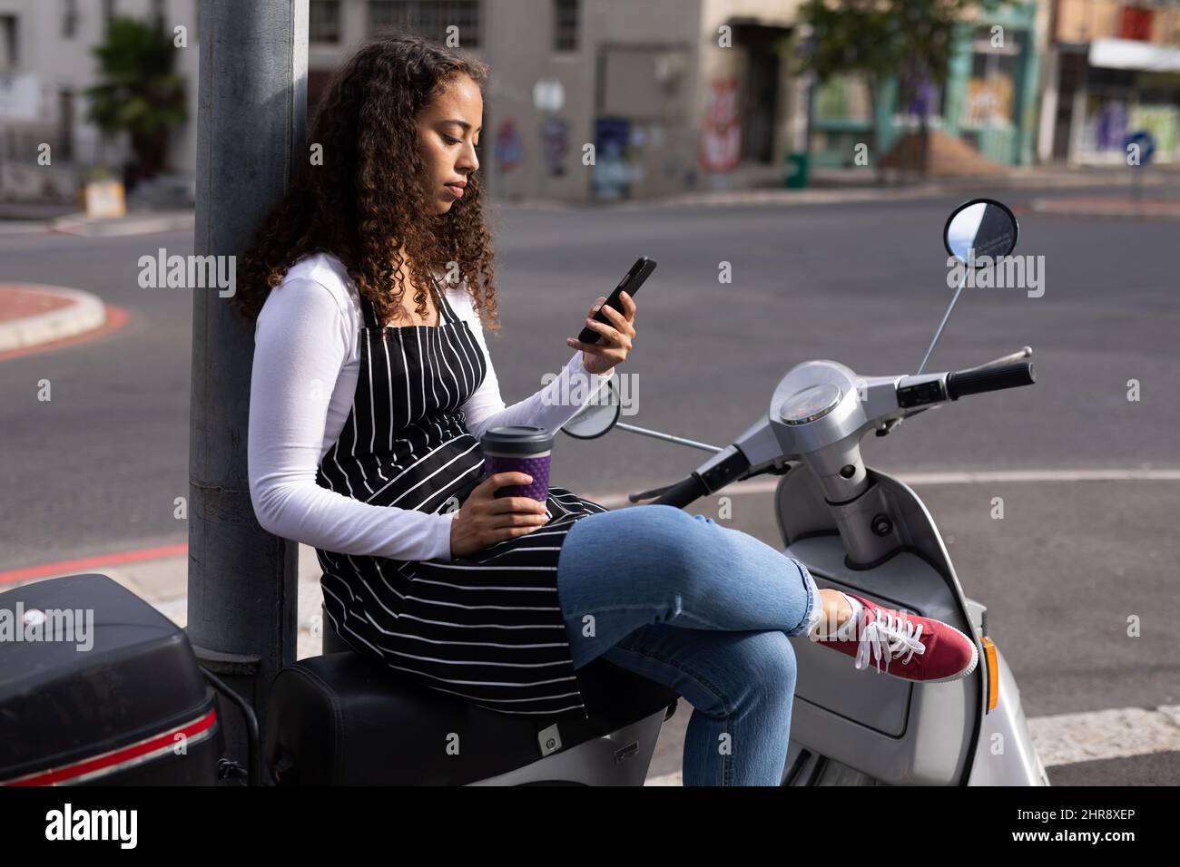 Giovane barista afroamericano che utilizza lo smartphone seduto con tazza monouso sullo scooter Foto Stock