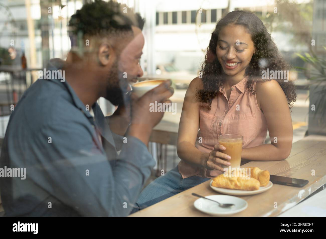 Giovane coppia afroamericana che parla mentre si siede con cibo e bevande in un caffè visto dalla finestra Foto Stock