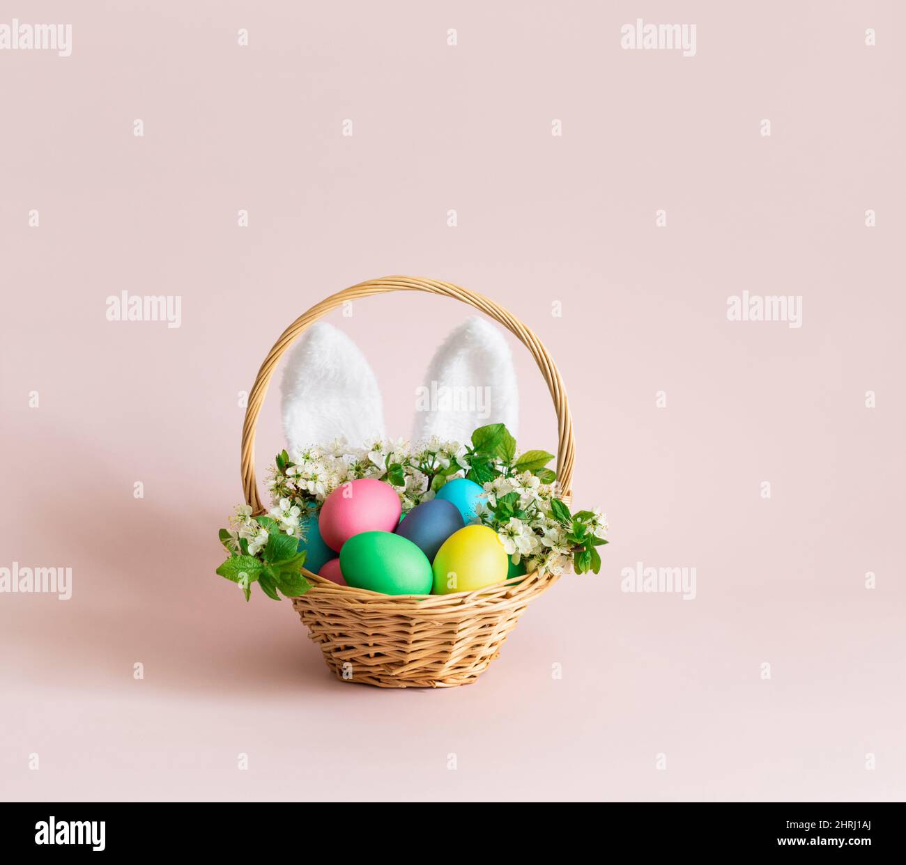 Uova di Pasqua con orecchie di coniglio e fiori in un cestino su sfondo rosa. Biglietto di auguri di Pasqua con spazio per il testo. Arredamento per le vacanze carino. Foto Stock