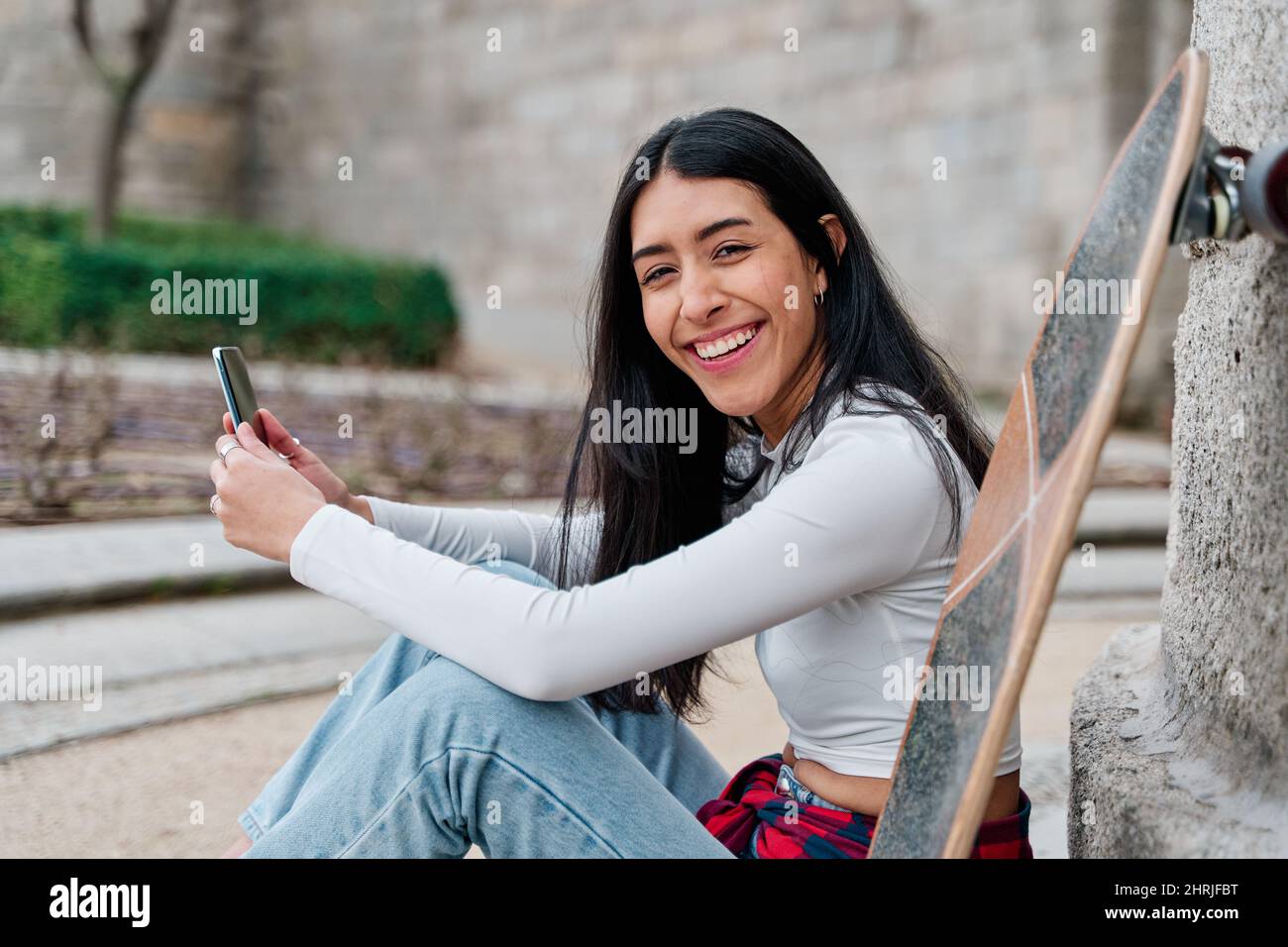 Giovane skater ragazza adolescente felice utilizzando uno smartphone guardando la fotocamera con sfondo sfocato Foto Stock