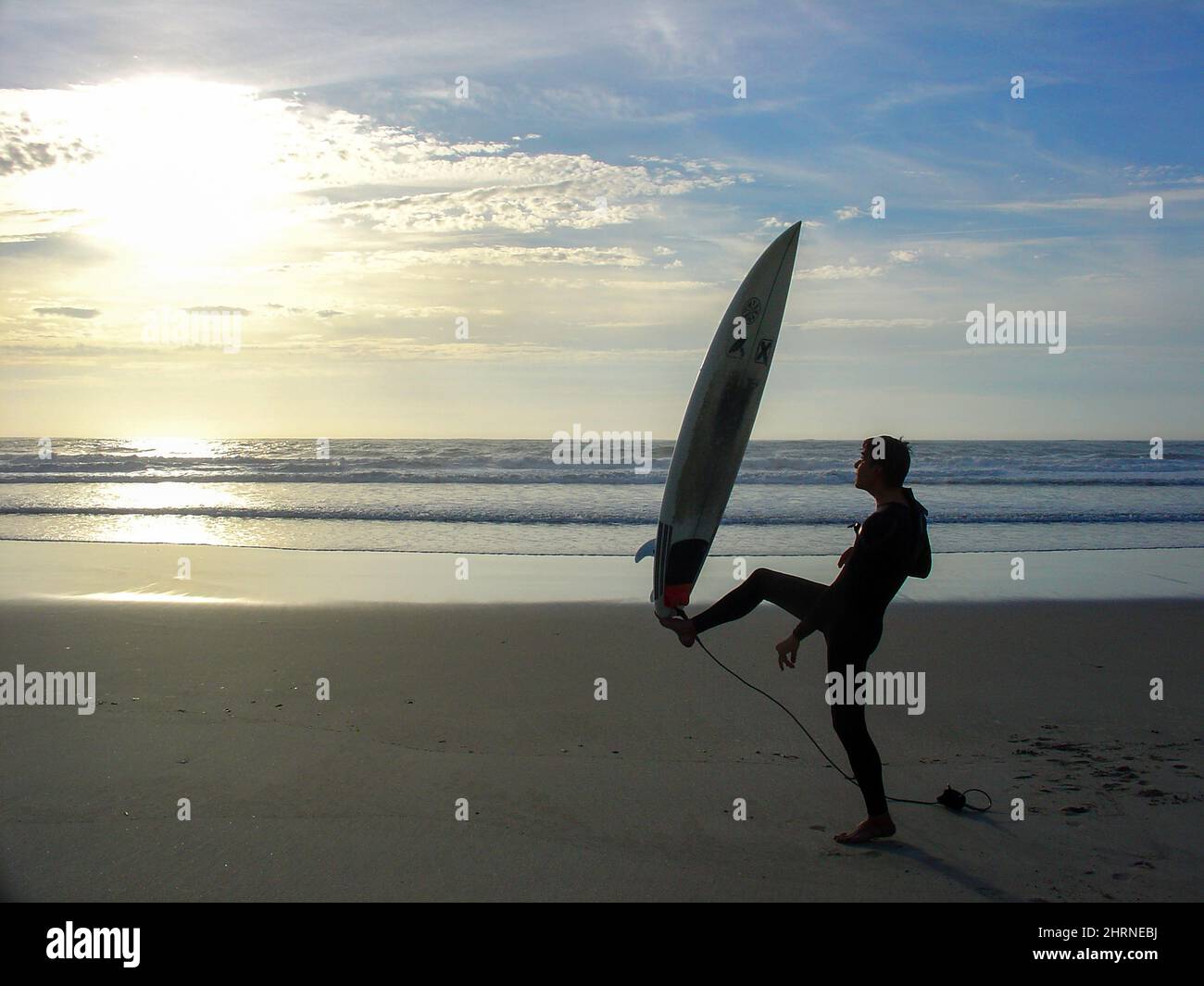 Surfer adulto maschio sul tramonto facendo esercizi o esponendo la sua tavola da surf. Surfista e yoga o stretching dopo una sessione di surf sulla spiaggia. Surf guru. Foto Stock