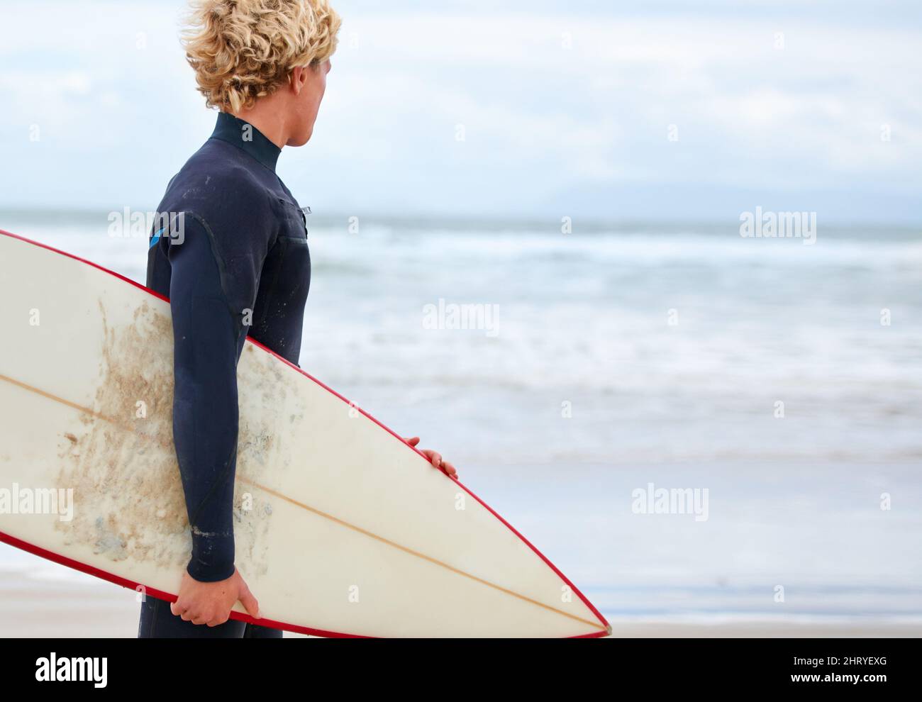 Alla ricerca di quella onda perfetta.... Un giovane surfista in piedi sulla spiaggia con l'oceano sullo sfondo. Foto Stock