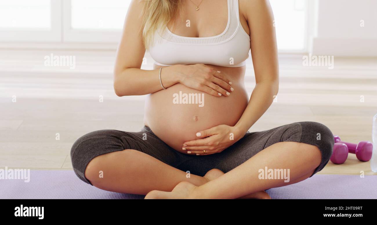 In buone mani. Scatto corto di una giovane donna incinta irriconoscibile strofinando il suo bump del bambino mentre si siede su un tappeto di yoga. Foto Stock
