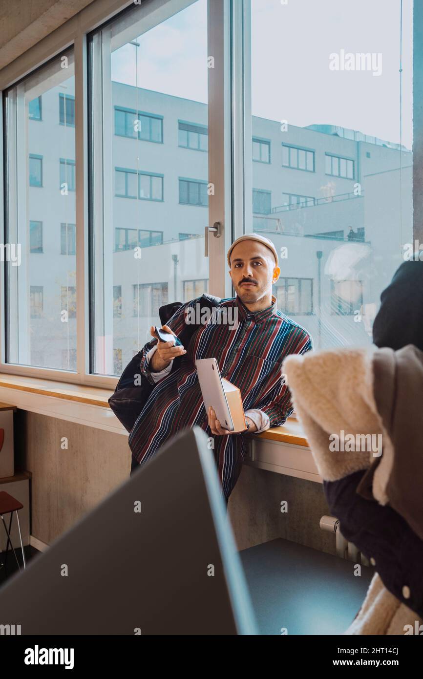 Ritratto di studente fidato maschio con smartphone e libro appoggiato sul davanzale in aula Foto Stock