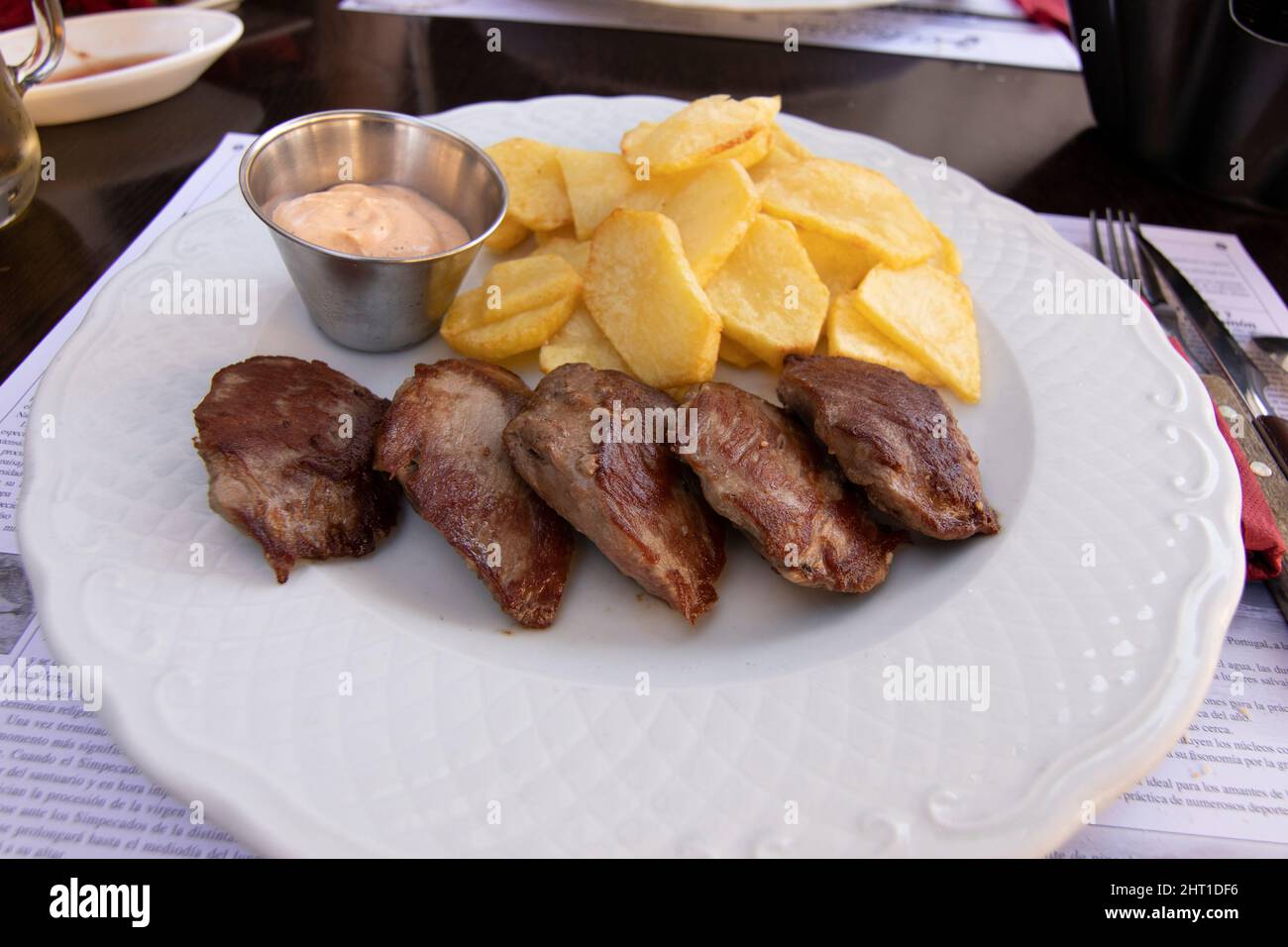 Bistecca di controfiletto alla griglia con patate a fette arrostite, il tutto su un piatto bianco. Una ciotola con salsa calda. Cucina spagnola. Foto Stock