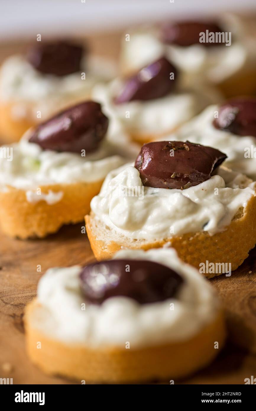 Molti piccoli pezzi di pane con tzatziki spalmato e frutta di oliva su tavola di legno cucina, cucina greca tradizionale. Primo piano. Foto Stock