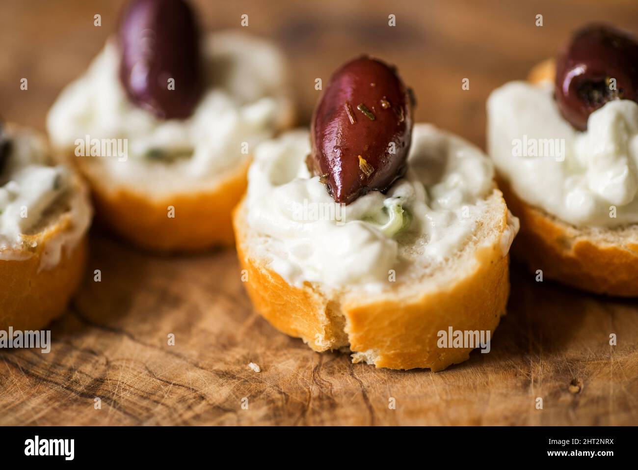 Piccolo pezzo di pane con tzatziki spalmato e frutta di oliva su tavola di legno da cucina di olivo, cucina greca tradizionale. Primo piano. Foto Stock