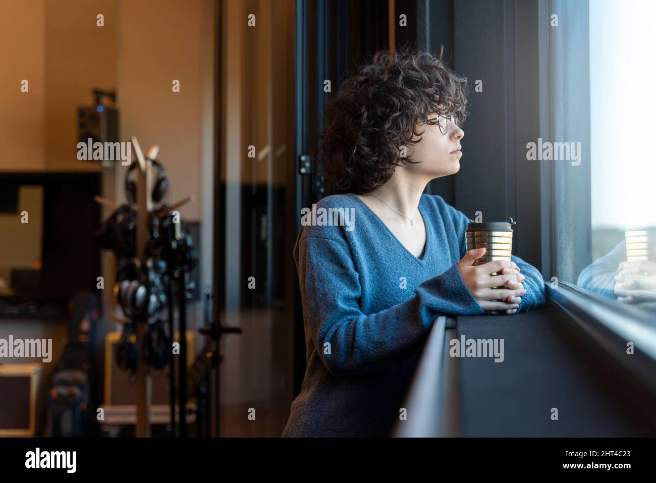 Una donna ricci capelli guarda fuori la finestra mentre tiene un vetro con la sua mano Foto Stock