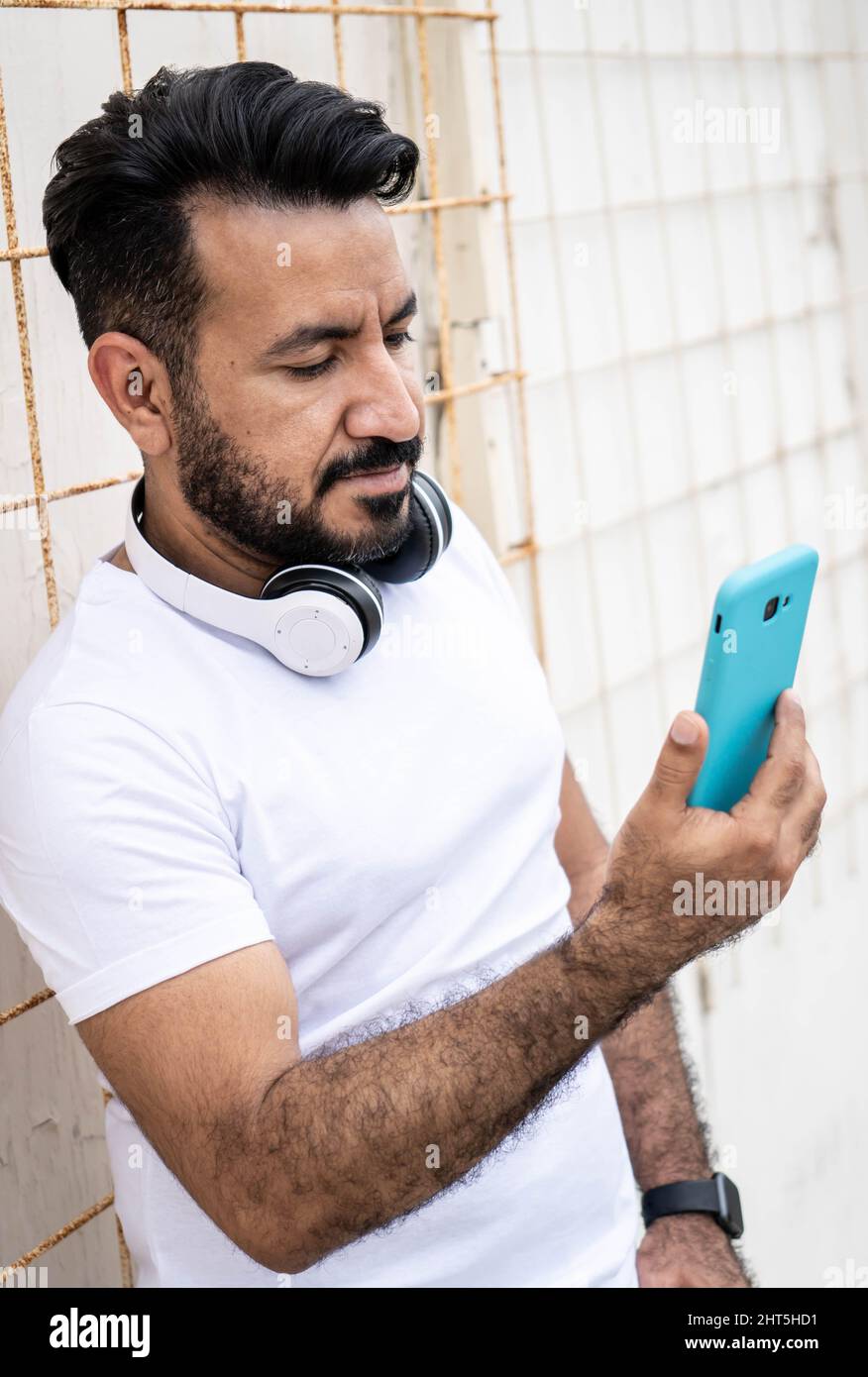 Uomo latino con una barba, guardando il suo cellulare appoggiato a un muro Foto Stock