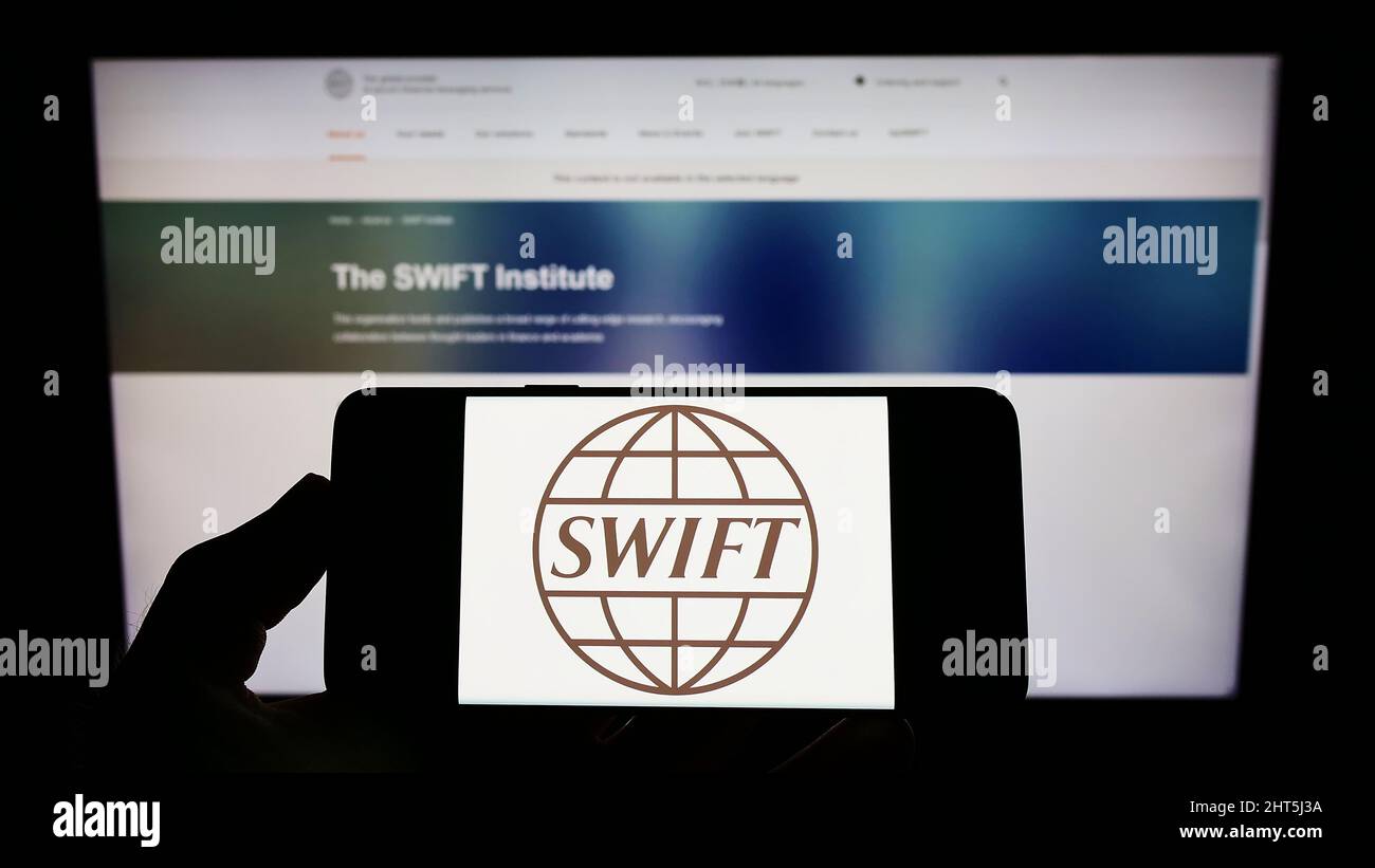 Persona che detiene il cellulare con il logo del sistema di transazioni bancarie SWIFT sullo schermo di fronte alla pagina web. Mettere a fuoco sul display del telefono. Foto Stock