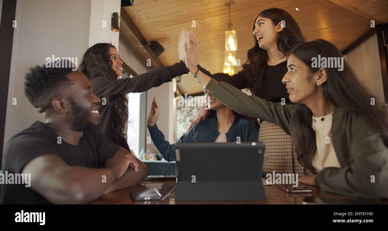 Gruppo di giovani dell'Asia del Sud che fanno i fives alti seduti ad un caffè con un tablet sul tavolo Foto Stock