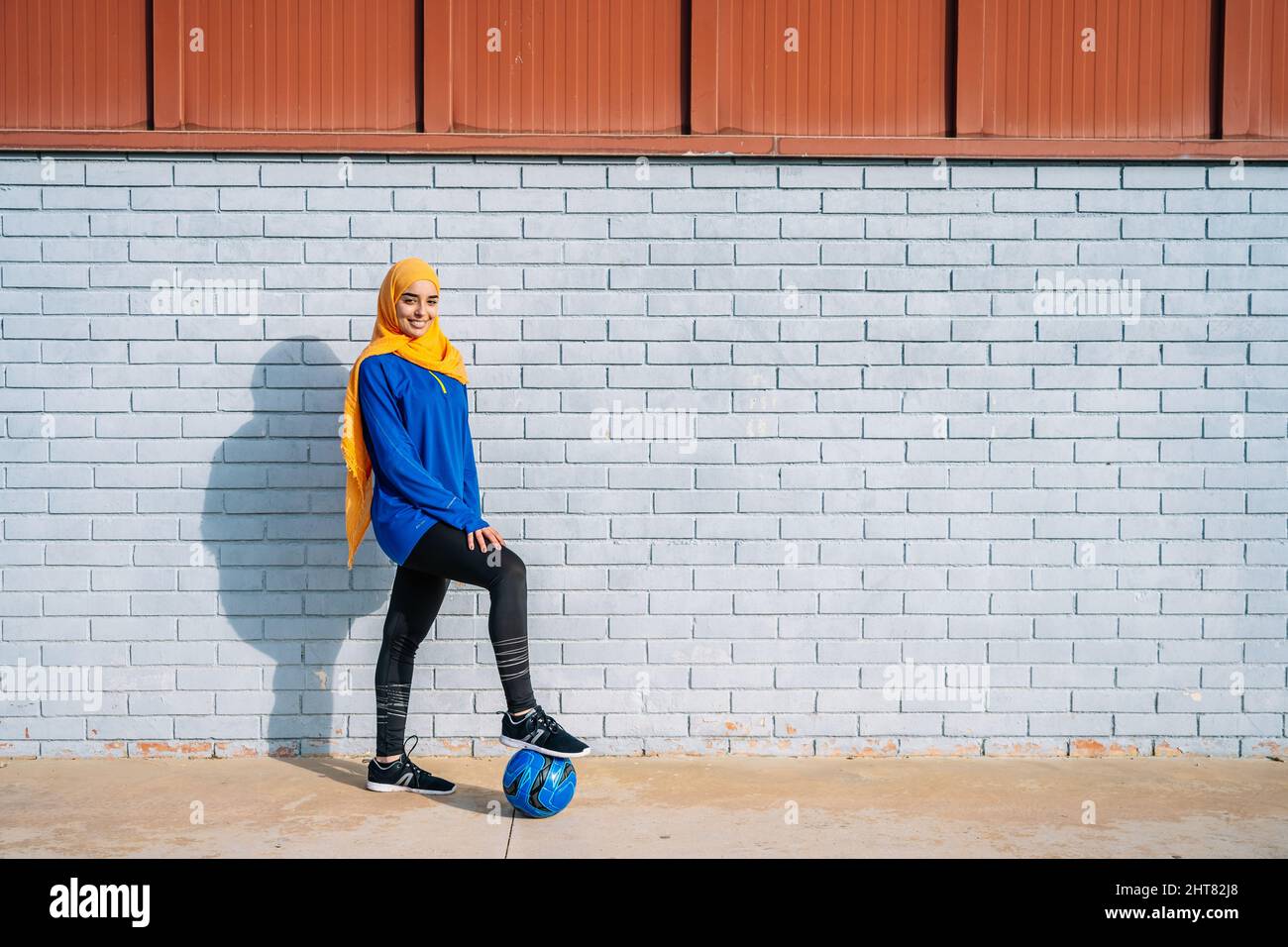 Un calciatore etnico sorridente con la gamba sulla palla vicino al muro di mattoni Foto Stock