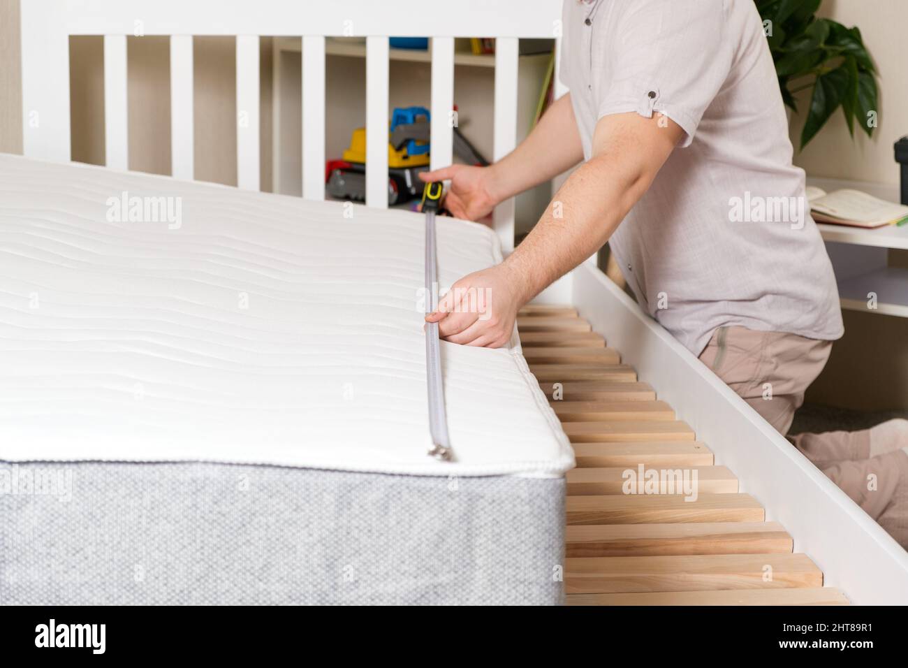 Un uomo misura la lunghezza del materasso del bambino per montarlo su un letto bianco di legno. Discrepanza tra le dimensioni del letto e del materasso. Foto Stock