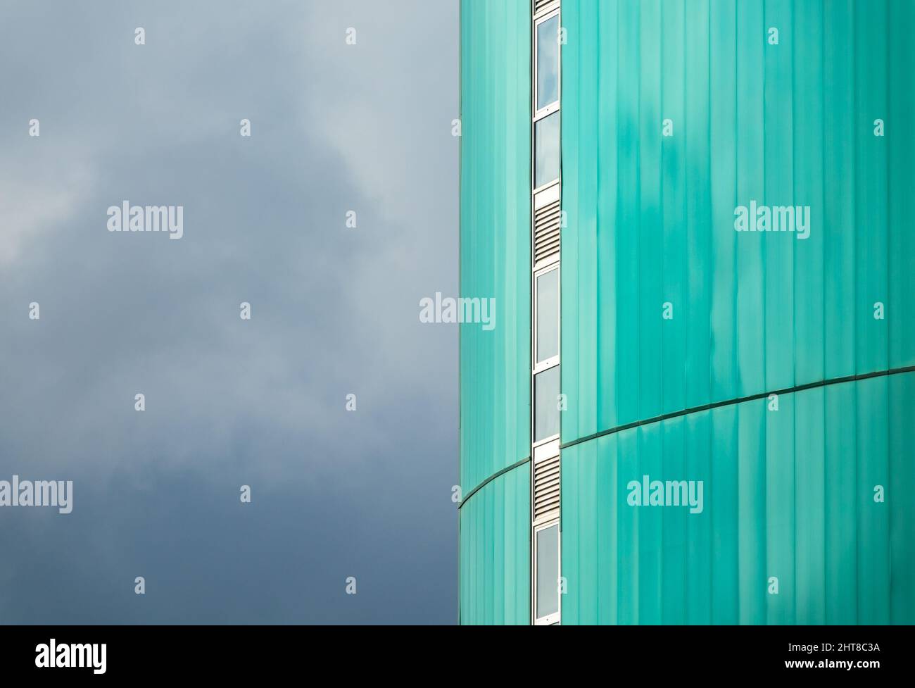 Vista della facciata verde di un alto edificio moderno con una fila verticale di finestre strette e il cielo nuvoloso sulla sinistra Foto Stock