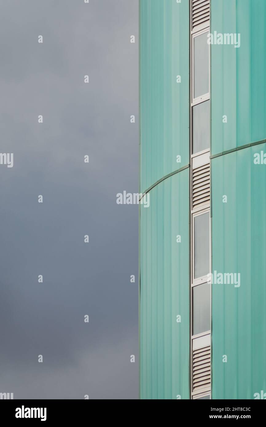 Vista della facciata verde di un alto edificio moderno con una fila verticale di finestre strette e il cielo nuvoloso sulla sinistra Foto Stock