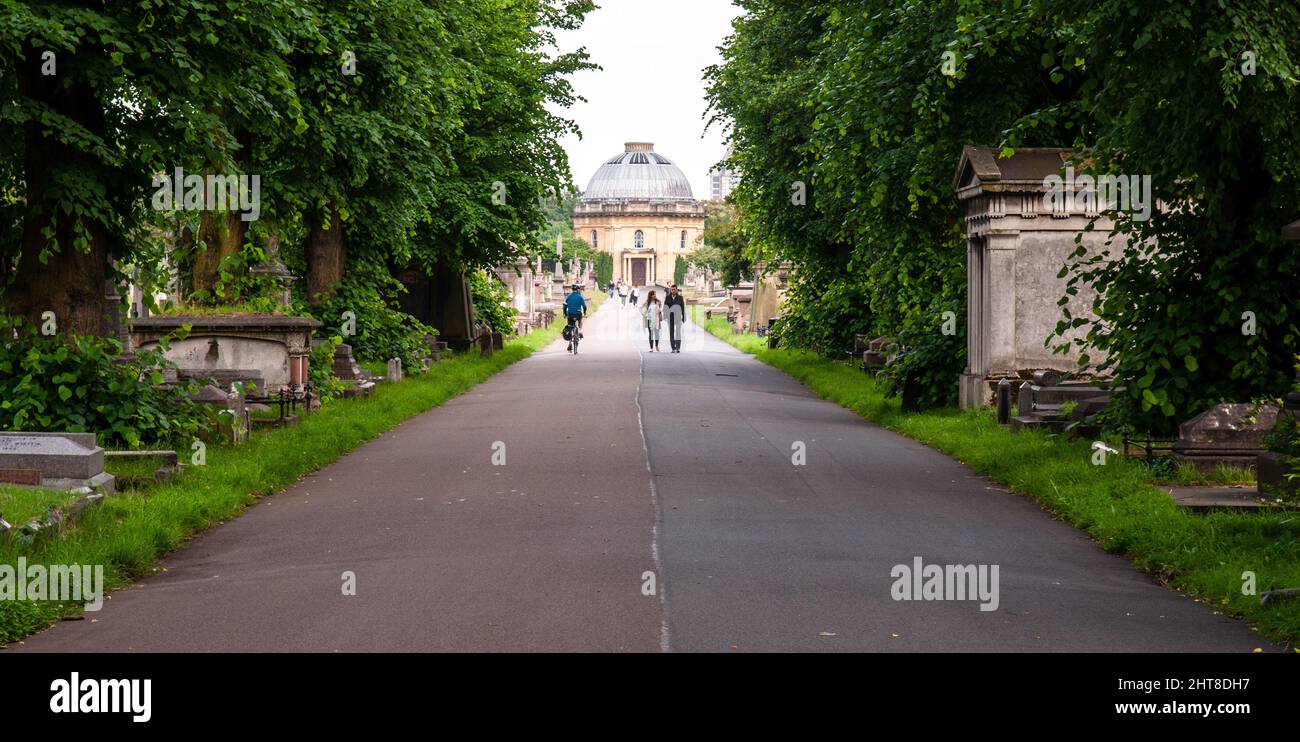 Londra, Inghilterra, Regno Unito - 18 giugno 2013: Pedoni e un ciclista passano tombe e lapidi su un sentiero attraverso il cimitero di Brompton a Chelsea, Londra. Foto Stock