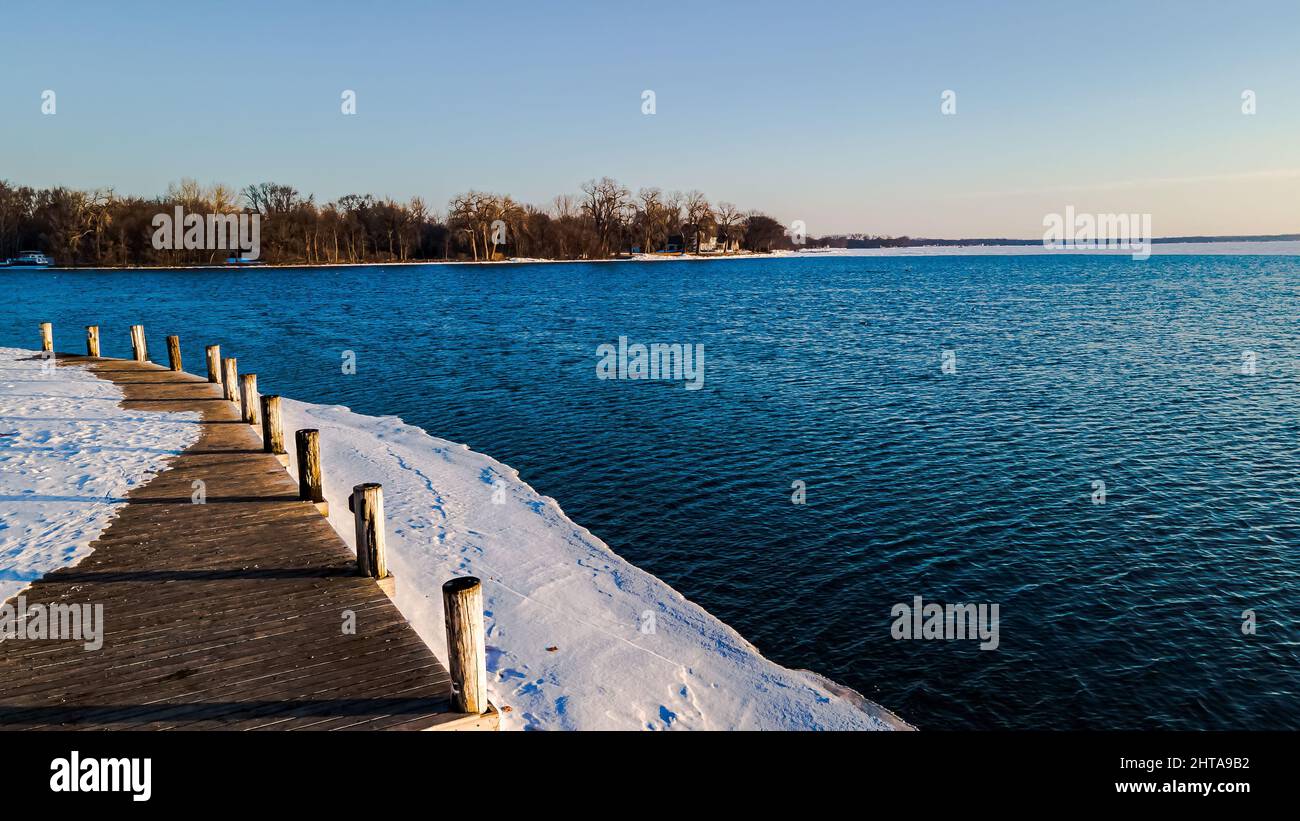 Mattina presto sul molo di legno con la neve che lo ricopre e il bordo ghiacciato dell'acqua. Foto Stock