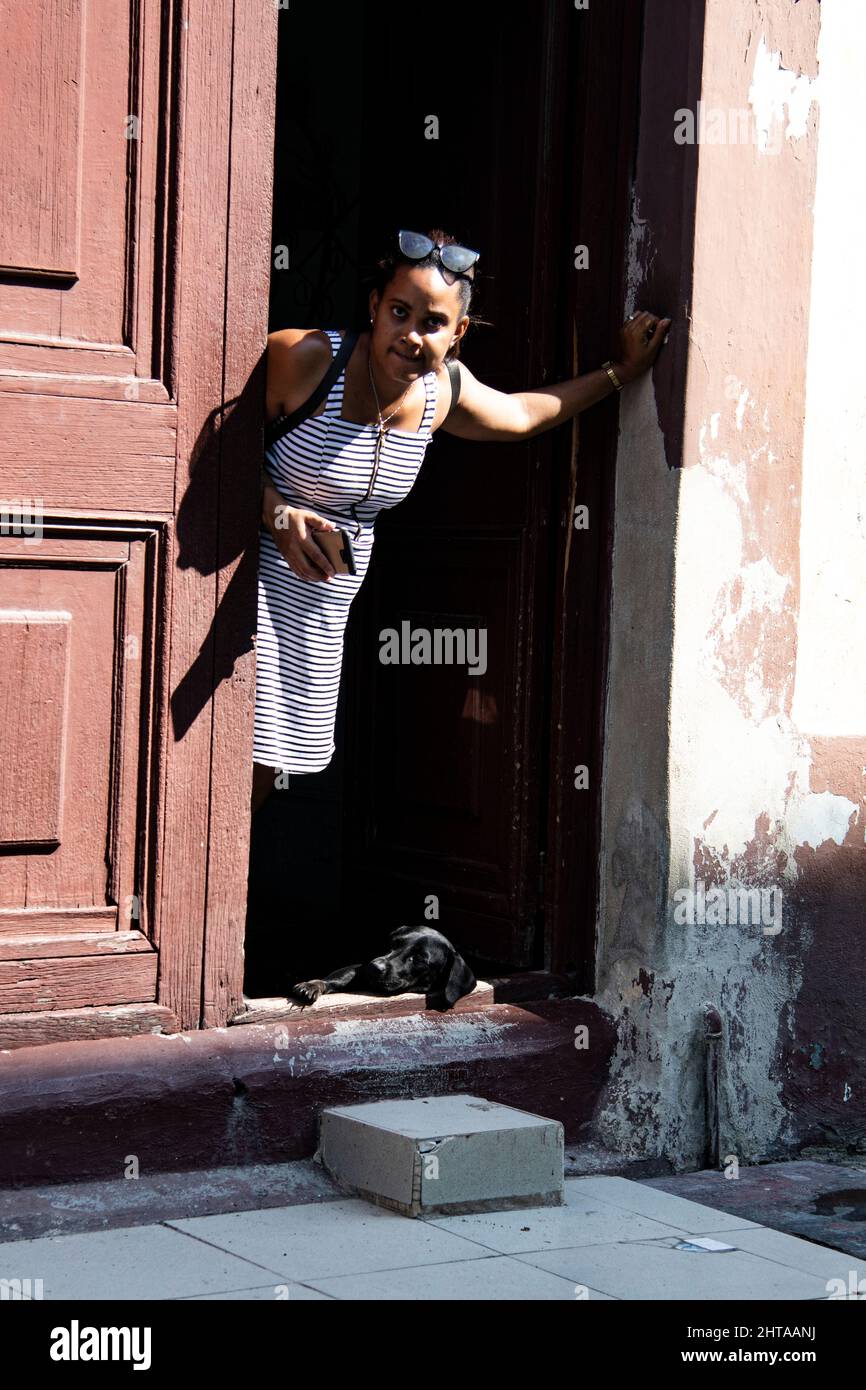 Una giovane donna tiene il suo cellulare e si appoggia contro la porta mentre il suo cane da compagnia si stende a terra nella porta della loro casa a l'Avana, Cuba. Foto Stock