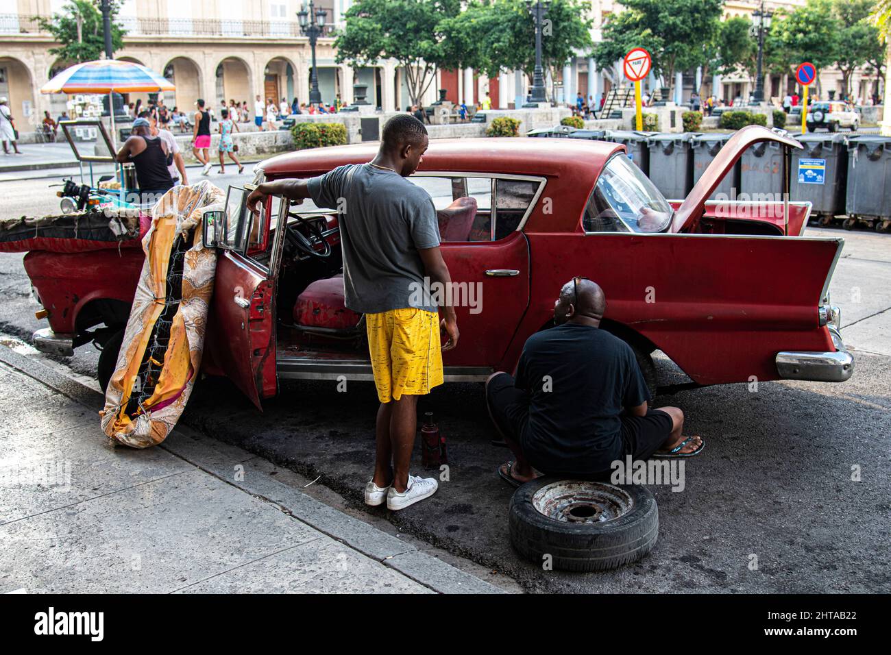 Due uomini cubani parlano mentre lavorano per fissare un pneumatico su una macchina a l'Avana, Cuba con un materasso sul cofano di una vecchia auto classica americana. Foto Stock