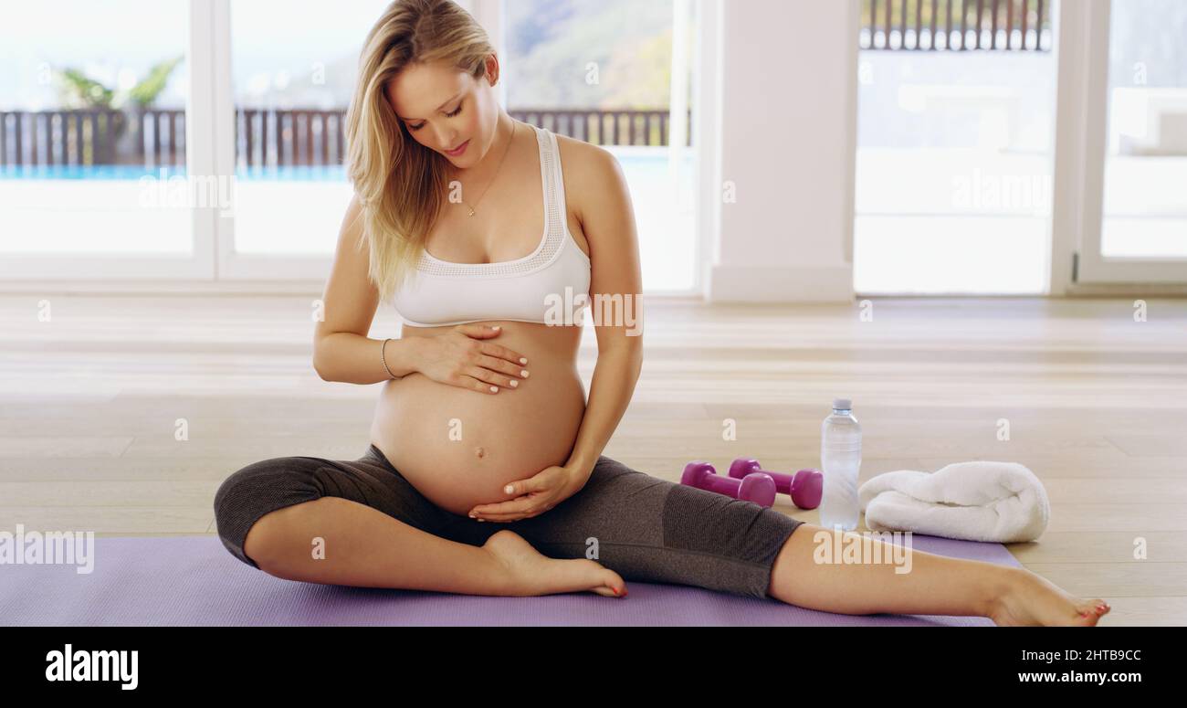 Non vedo l'ora di incontrarti. Scatto corto di una giovane donna incinta attraente che sfrega il suo bump del bambino mentre si siede su un tappeto di yoga. Foto Stock