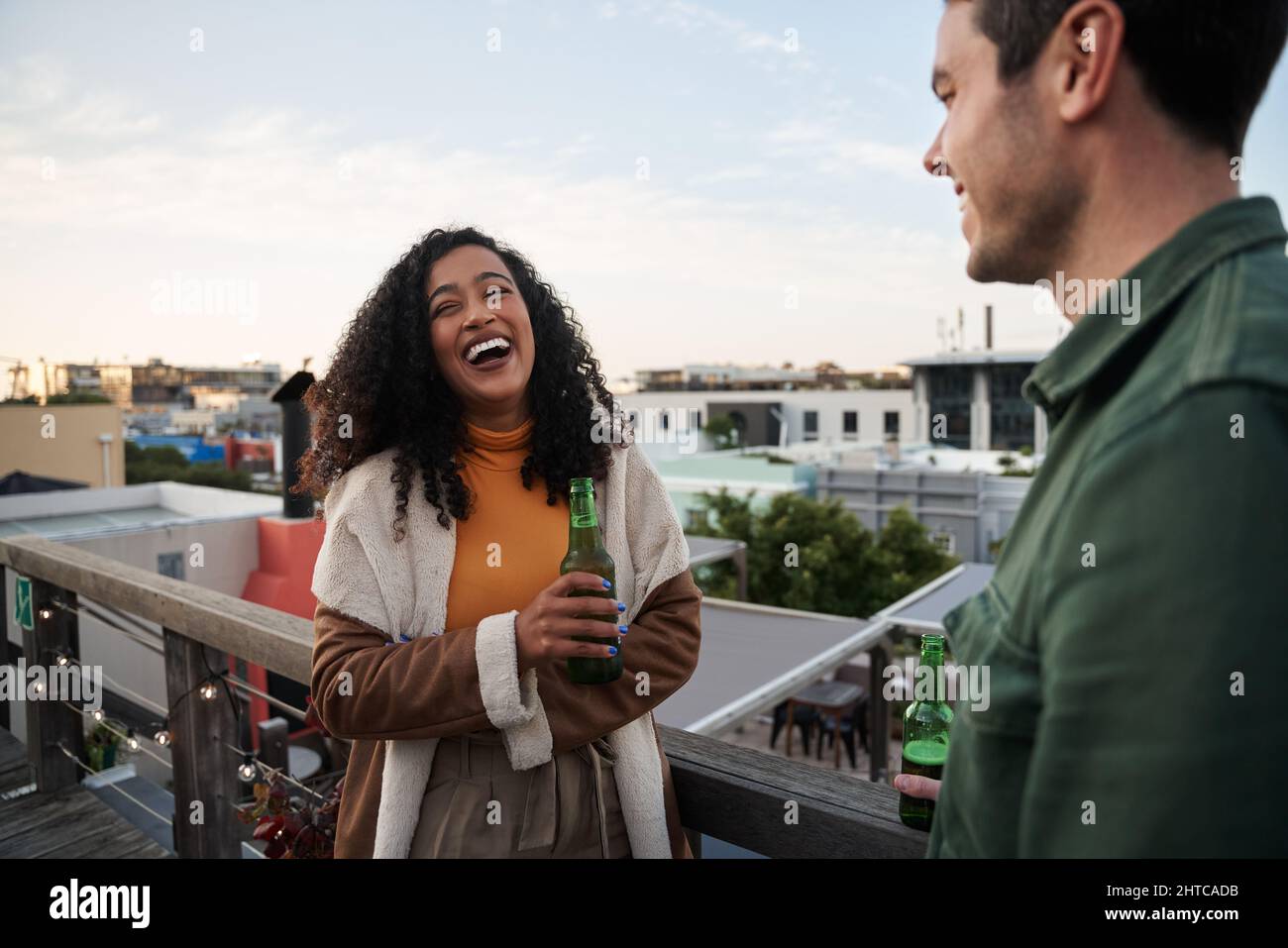 Biracial giovane adulta femmina ridendo con un amico sul balcone di una terrazza sul tetto della città. Drink a portata di mano, vista sulla città Foto Stock