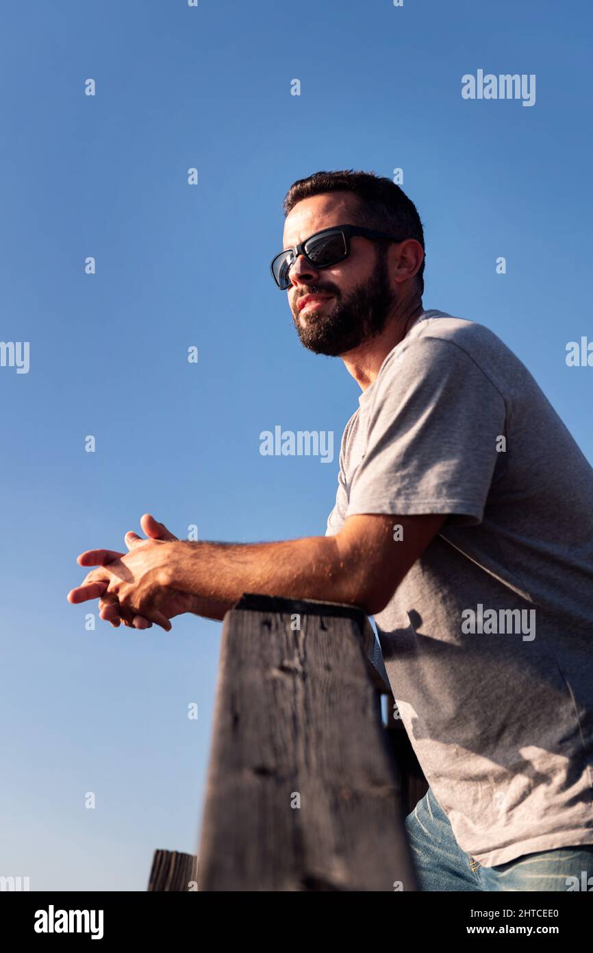ritratto verticale di un bel giovane uomo con occhiali da sole appoggiati su una ringhiera di legno con cielo blu sullo sfondo, concetto di tempo libero e relax, c Foto Stock