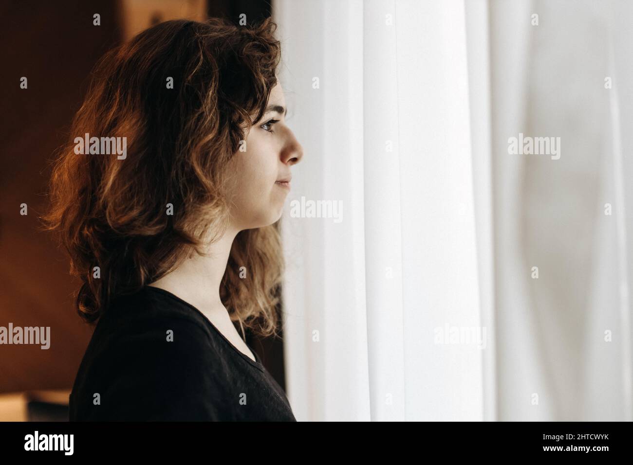 Giovane ragazza pensierosa con capelli ricci che guardano alla finestra Foto Stock