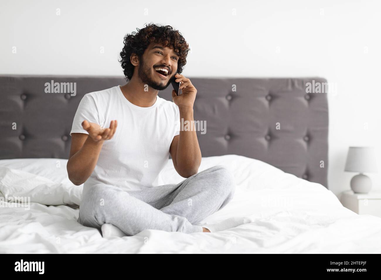 Un ragazzo indù positivo seduto a letto, che parla al telefono Foto Stock