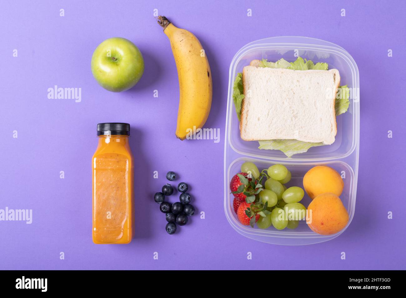 Vista dall'alto di frutta e bottiglia di succo di frutta in sandwich in scatola tiffin su sfondo viola Foto Stock