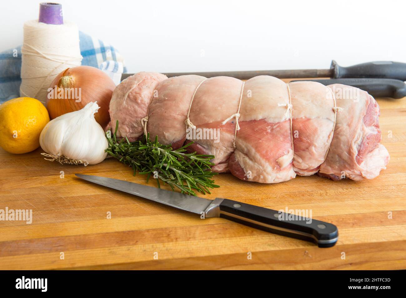 Spalla cruda disossata e arrotolata di agnello preparata per la tostatura su un tagliere di legno con un filo da cucina a rullo un coltello rosmarino limone e cipolla Foto Stock