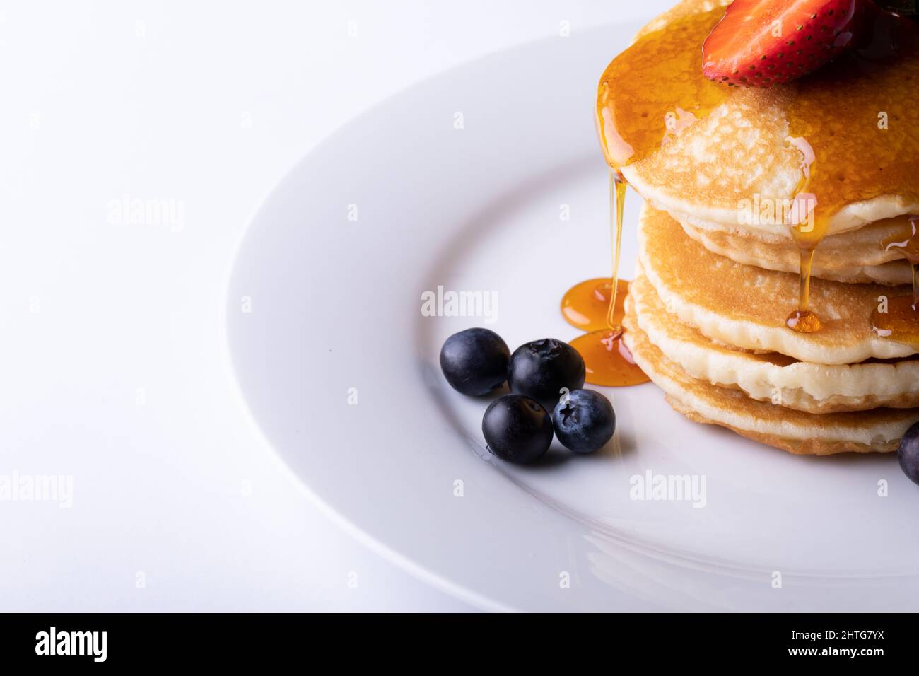 Primo piano dello sciroppo che gocciola sopra le frittelle accatastate con la frutta della bacca nel piatto su sfondo bianco Foto Stock