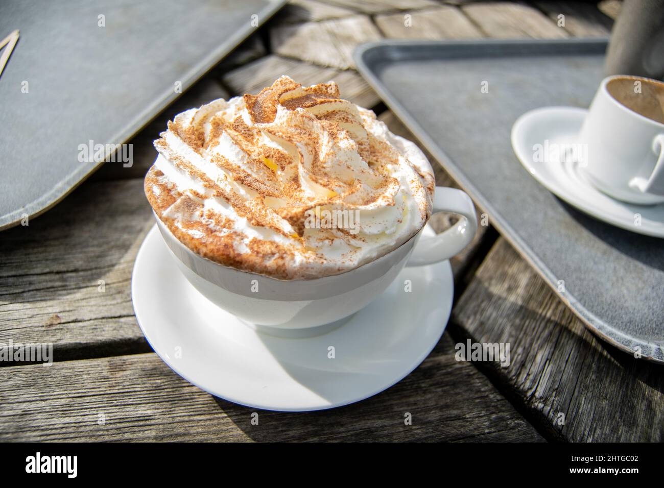 Primo piano per una grande tazza bianca di cioccolata calda con panna montata e spruzzi di cacao sulla parte superiore, tazza con bevanda standig sul tavolo esterno in legno accanto a. Foto Stock Primo piano per una grande tazza bianca di cioccolata calda con panna montata e spruzzi di cacao sulla parte superiore, tazza con bevanda standig sul tavolo esterno in legno accanto a. Foto Stock