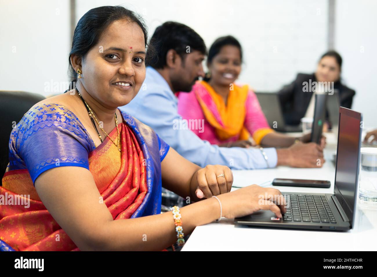 Sorridente donna indiana che guarda la macchina fotografica mentre lavora sul laptop in ufficio - concetto di empowerment donna, emozione positiva e sviluppo Foto Stock