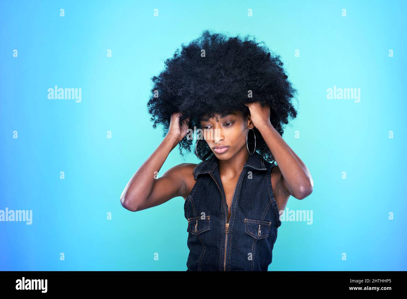 Elegante donna afroamericana in abito in denim che mantiene le mani in capelli ricci su sfondo blu in Vivid studio Foto Stock