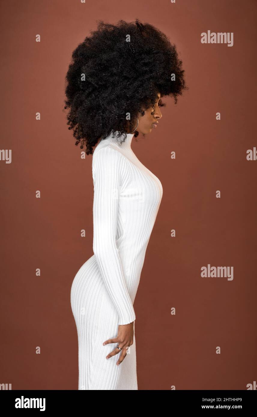 Vista laterale del corpo modello femminile afroamericano con acconciatura nera afroamericana indossando elegante abito bianco su sfondo marrone in studio Foto Stock