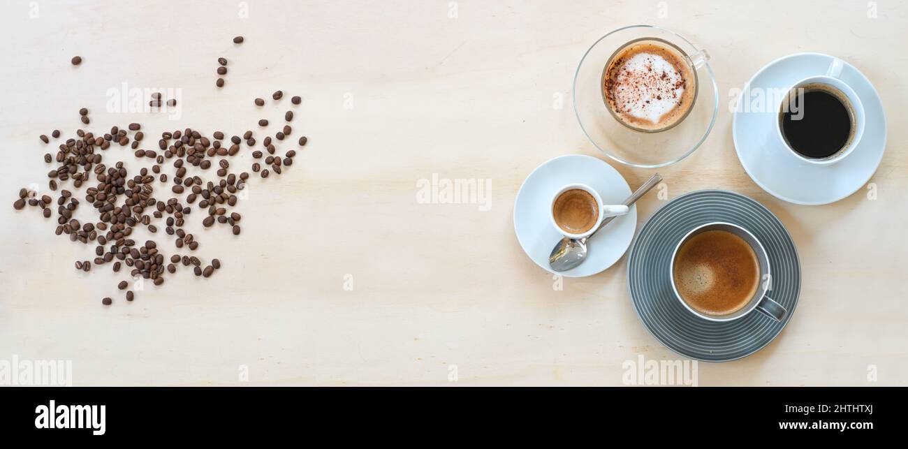 Caffè in chicchi arrosto e quattro tazze con varie bevande come espresso, cappuccino, nero lungo e con crema su un tavolo di legno chiaro, forma panoramica Foto Stock