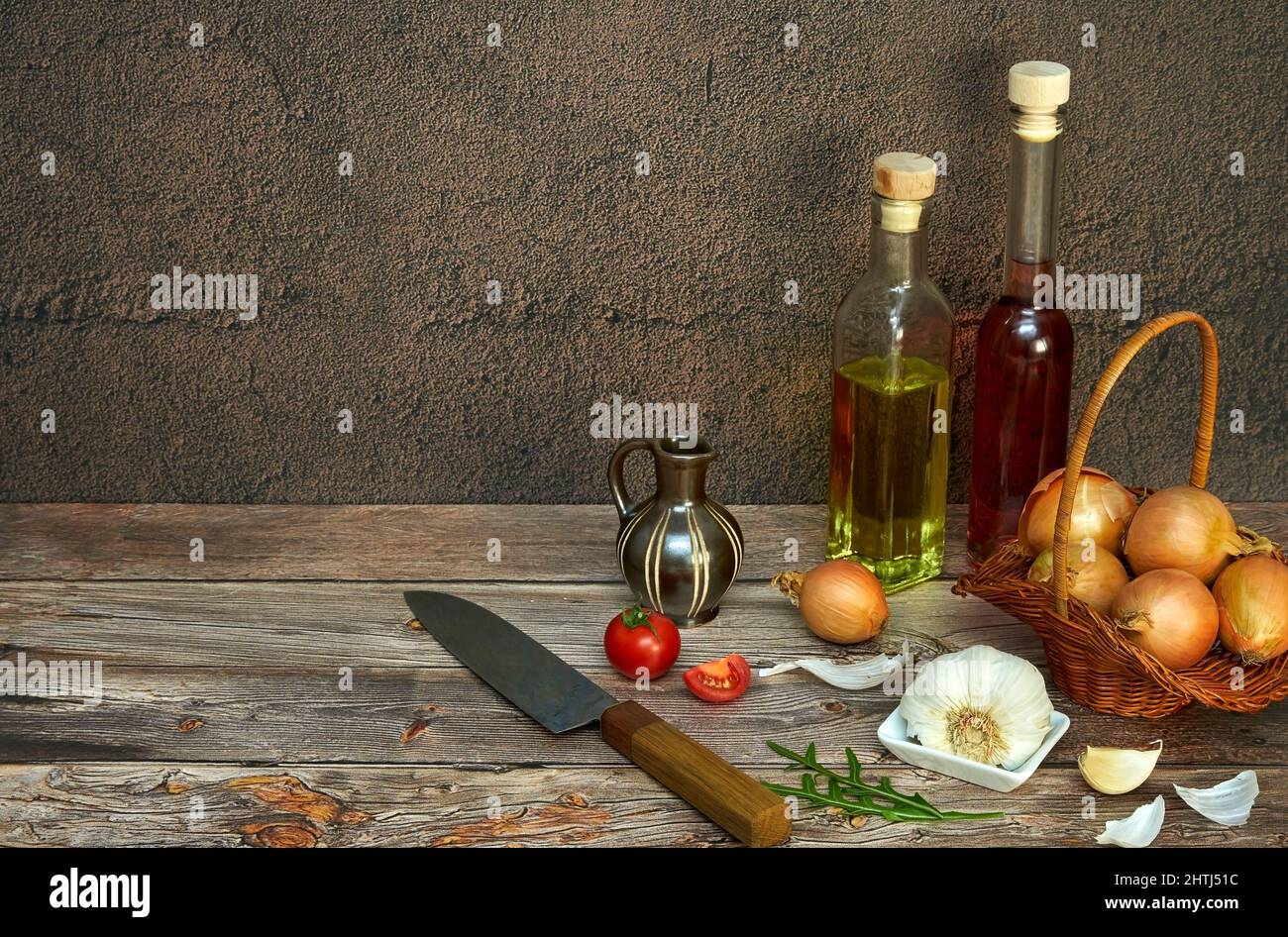 Küchenmotiv mit Zwiebel in einem Körbchen, Knoblauch, Tomaten, Rucola, Fruchtessig, Und Öl mit Messer auf Holzuntergrund Foto Stock