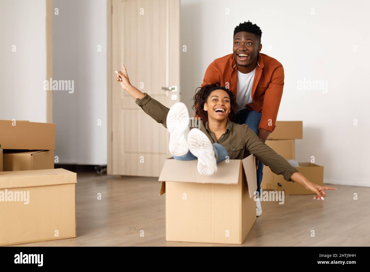 Happy African Man Riding Woman in Moving Box in Home Foto Stock