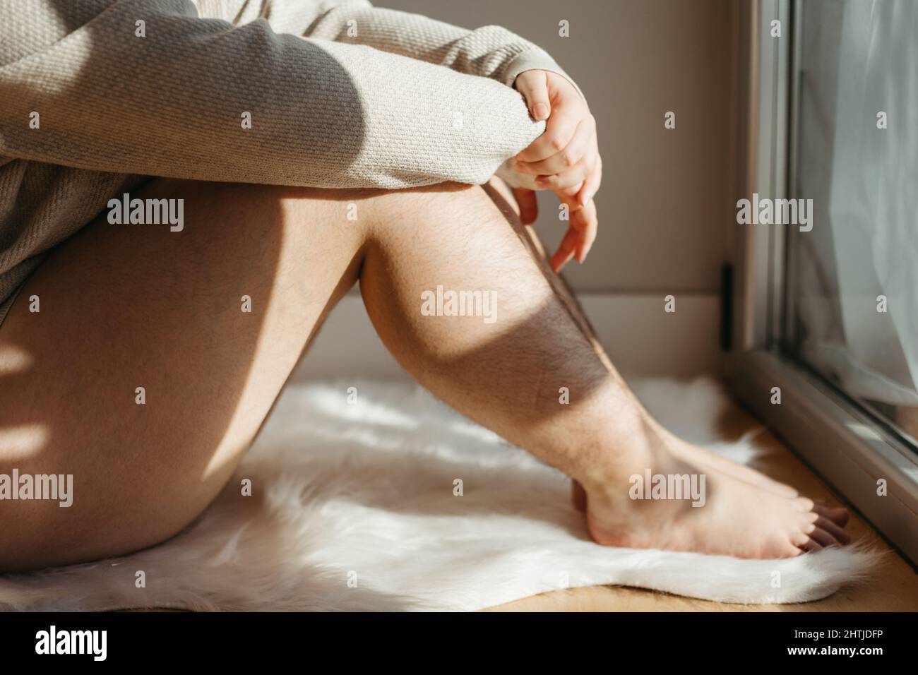 Vista laterale di femmina non riconoscibile dal taglio, con gambe pelose sedute vicino alla parete e finestre a casa mentre si gode il tempo Foto Stock
