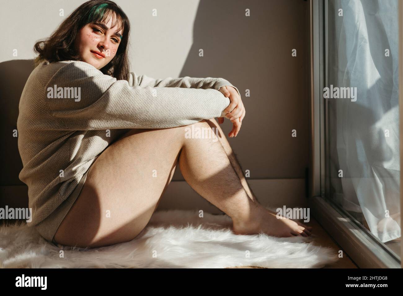Vista laterale della donna con gambe pelose sedute vicino alla parete e finestre che guardano la macchina fotografica a casa mentre si gode il tempo Foto Stock