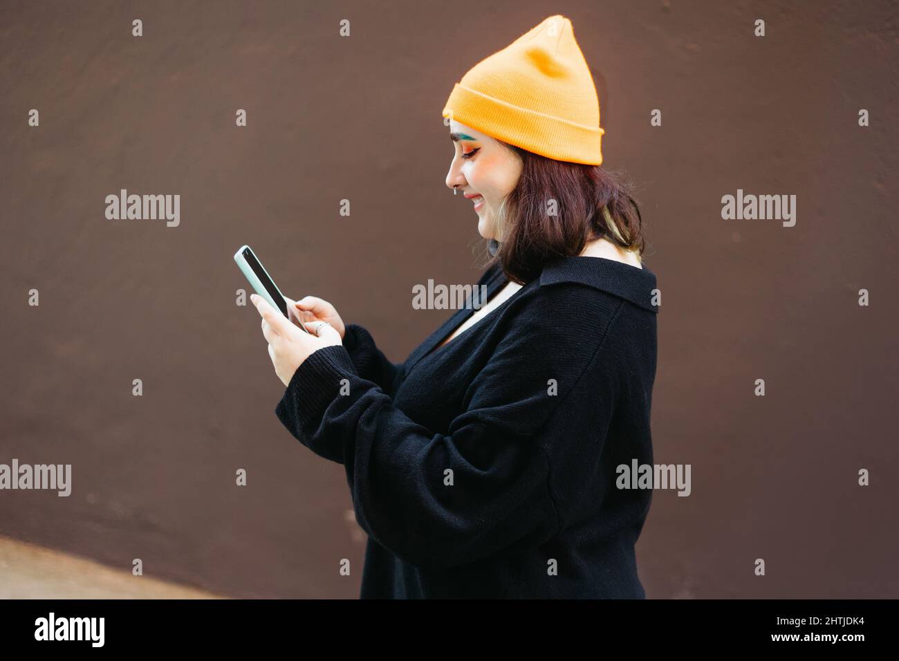 Vista laterale del contenuto femminile con trucco luminoso in cappello messaggi di testo sul cellulare mentre in piedi sulla strada contro sfondo marrone Foto Stock