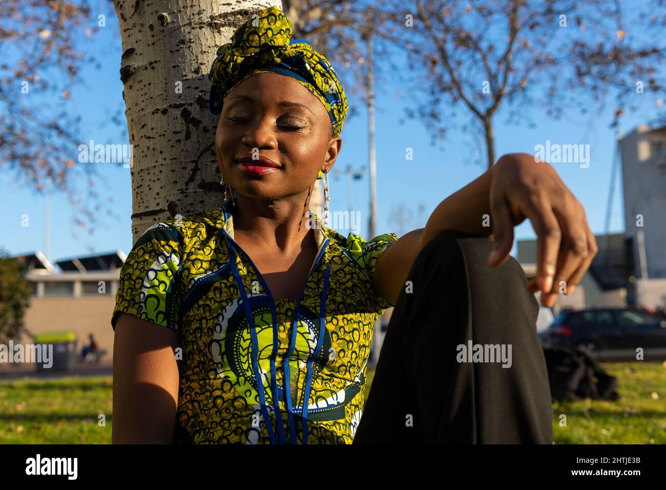 Donna nera in blusa tradizionale e foulard seduta vicino ad albero tronco con occhi chiusi in giornata di sole nel parco della città Foto Stock