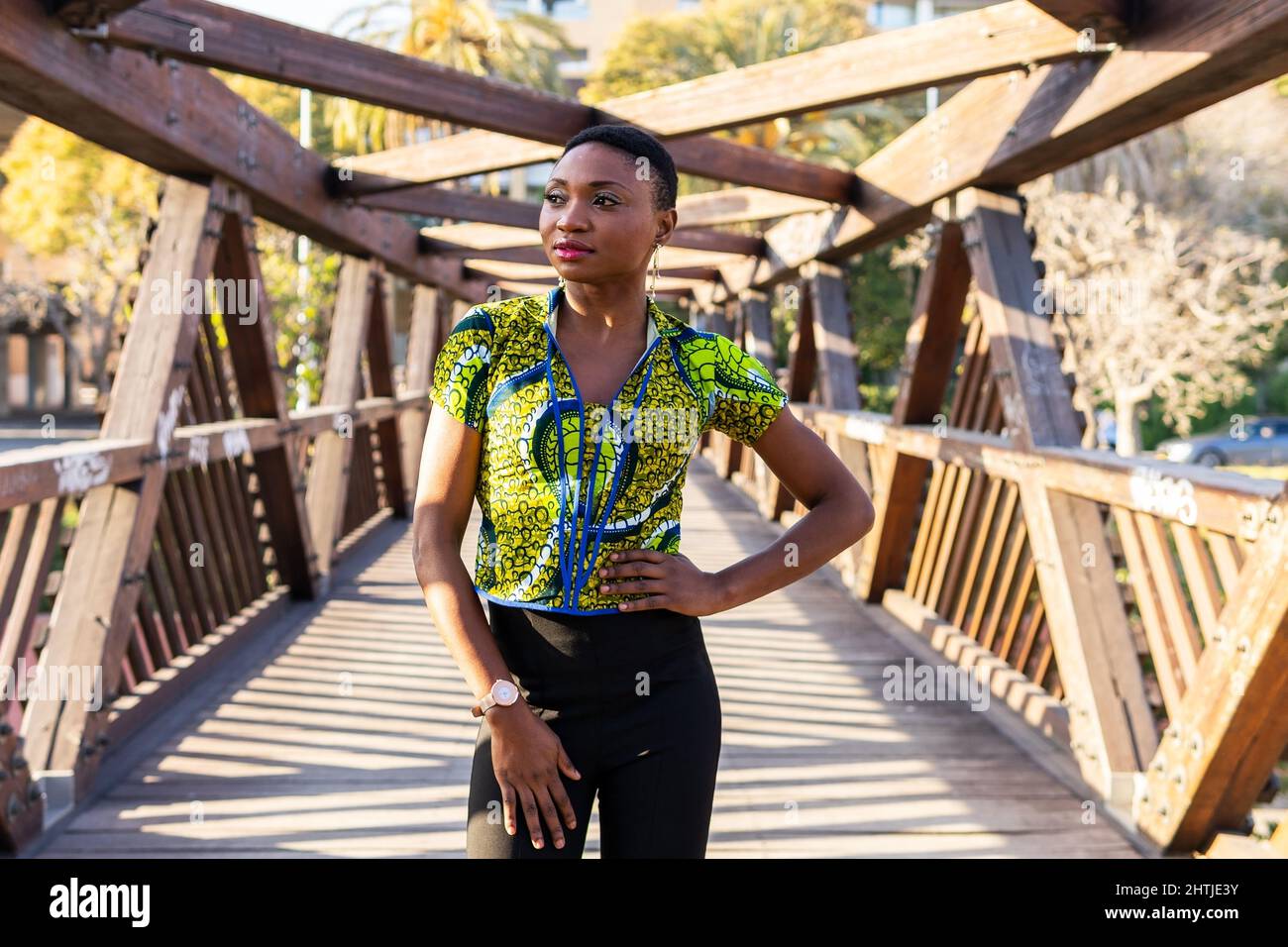 Donna Africana assicurata con i capelli corti che tengono la mano sulla vita e guardando via mentre si leva in piedi sul ponte del legname nella giornata di sole nel parco Foto Stock