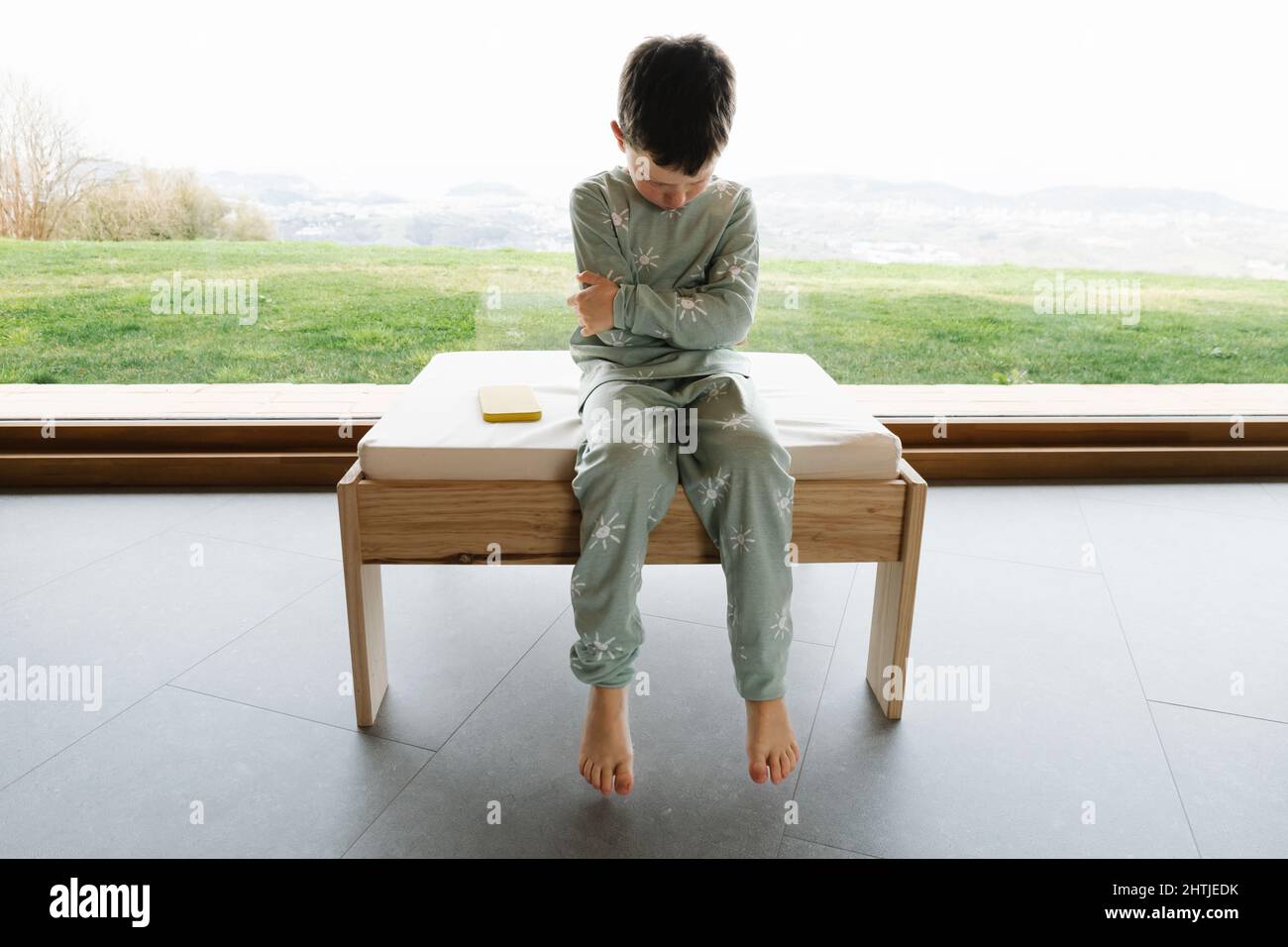 Ragazzo infelice in pajama seduto sul pouf in stanza luminosa il mattino vicino cellulare moderno a senza volto Foto Stock