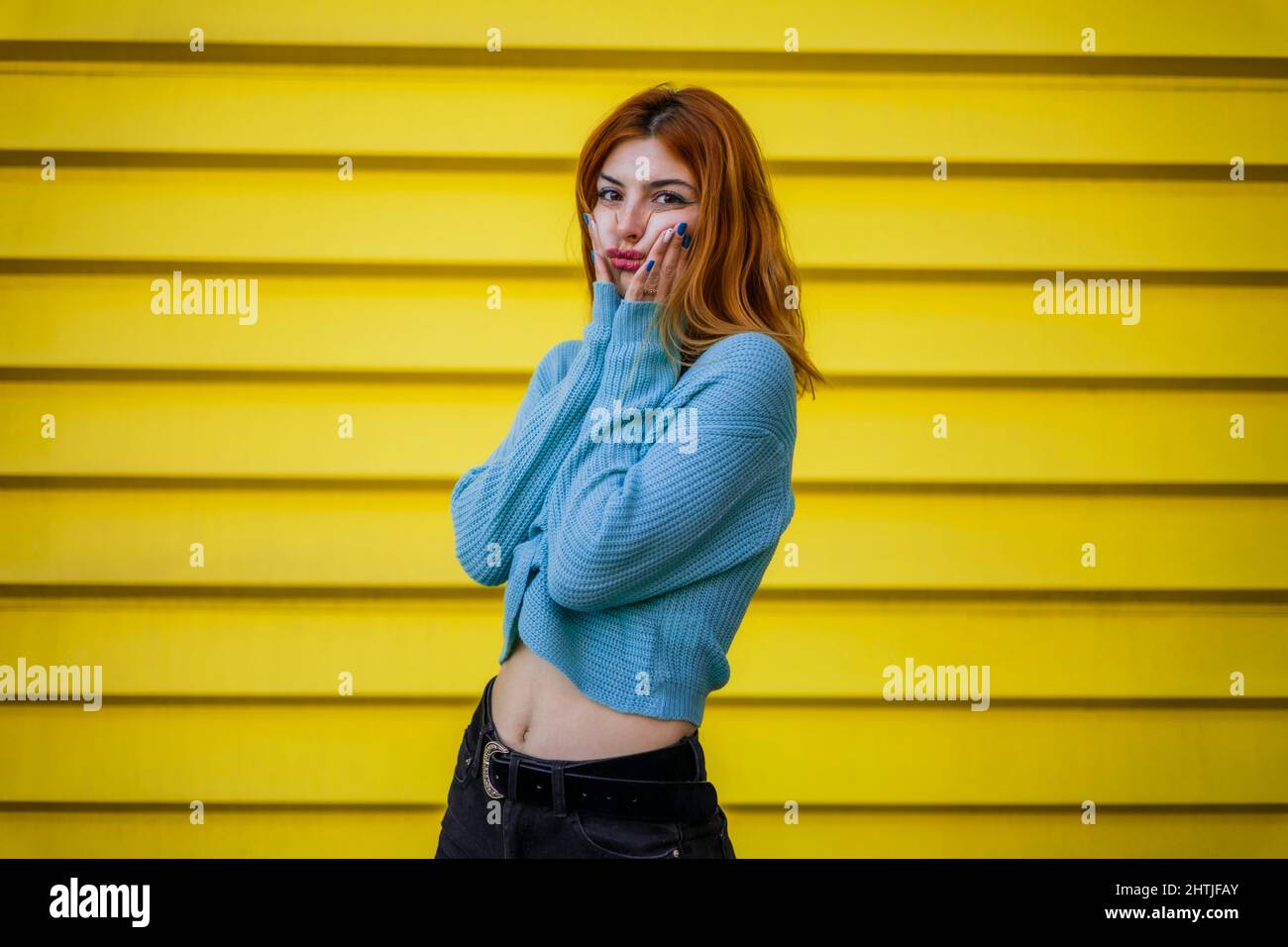 Donna premurosa che tocca le guance e guarda la macchina fotografica con il volto pensivo mentre si alza in piedi sulla strada su sfondo giallo luminoso in città Foto Stock