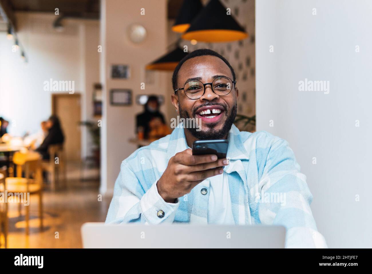 Happy afroamericano maschio guardare via e registrare audio messaggio sullo smartphone mentre si siede al tavolo con computer portatile in moderna caffetteria Foto Stock