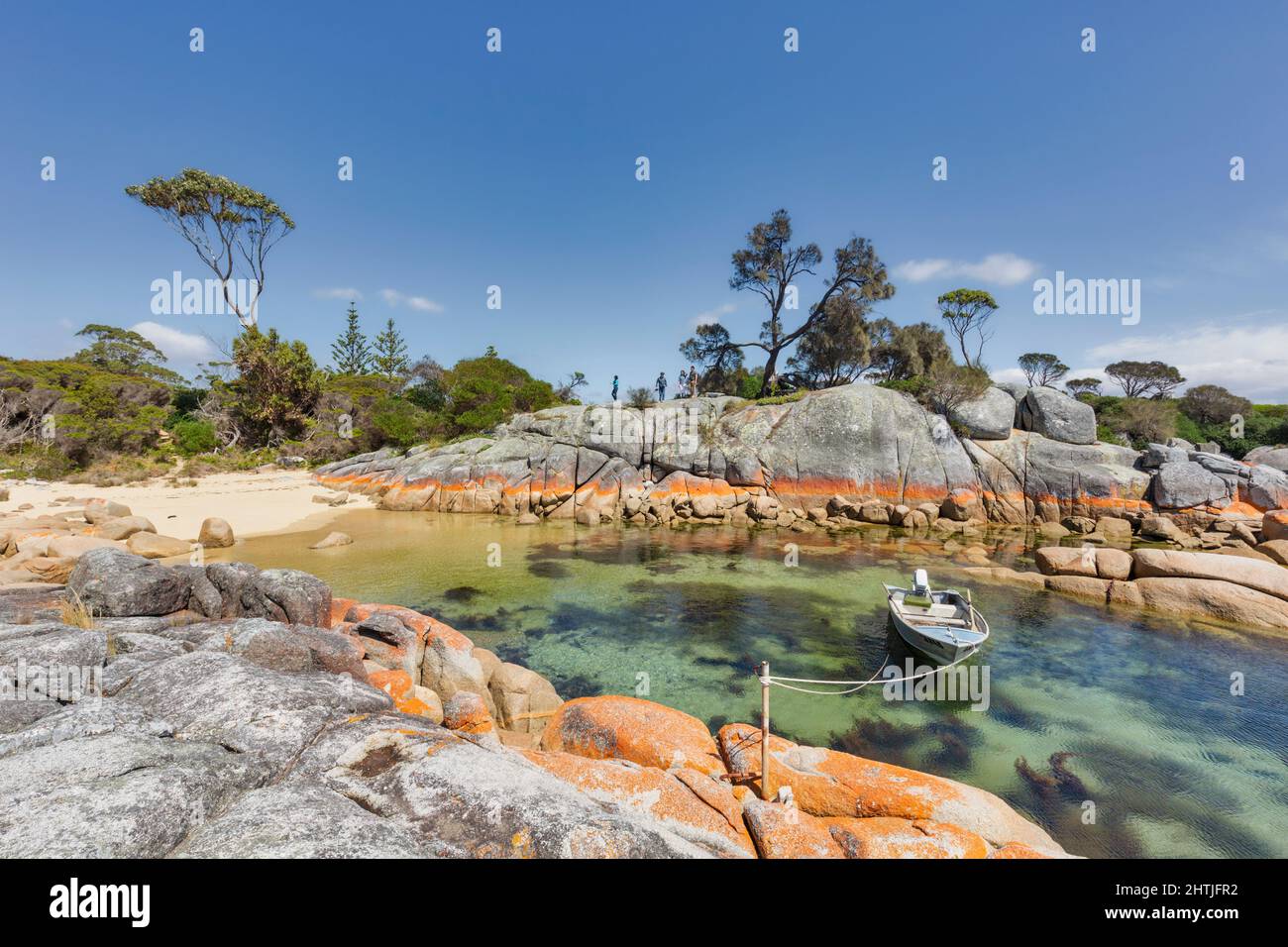 La crosta arancione del lichen Calapilla marina sulla roccia lungo la riva di Binalong Bay, Tasmania, Australia. Barca ormeggiata in baia. Foto Stock