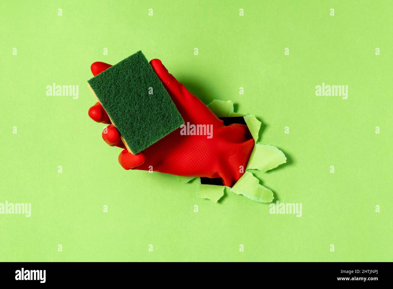 Una mano in un guanto di gomma con spugna per lavastoviglie fuoriesce da un foro lacerato di carta verde. Concetto di pulizia. Foto Stock