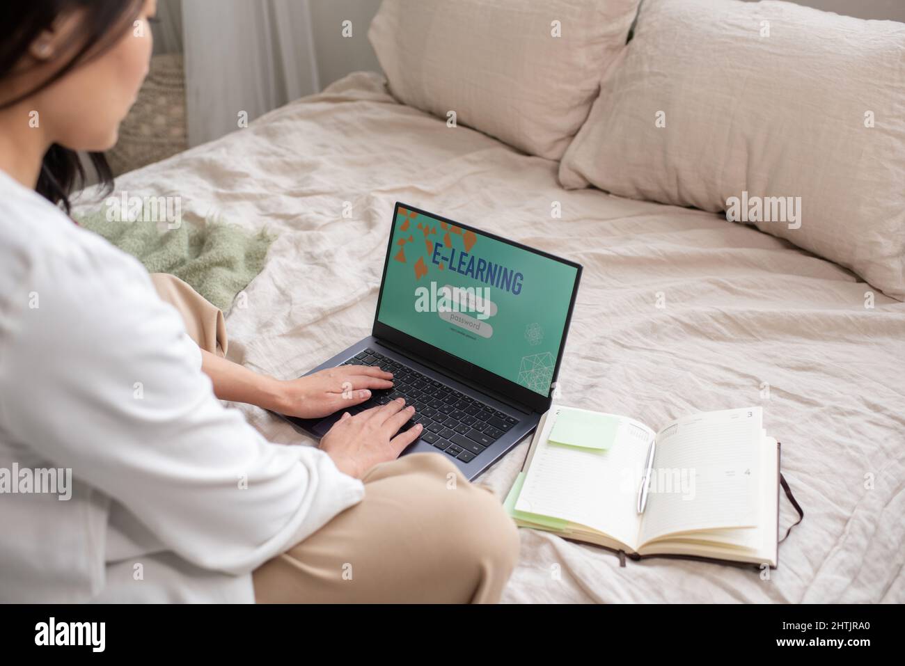 Giovane studentessa in casualwear seduta a letto e inserendo il suo nome e la password per accedere al corso di studio online Foto Stock