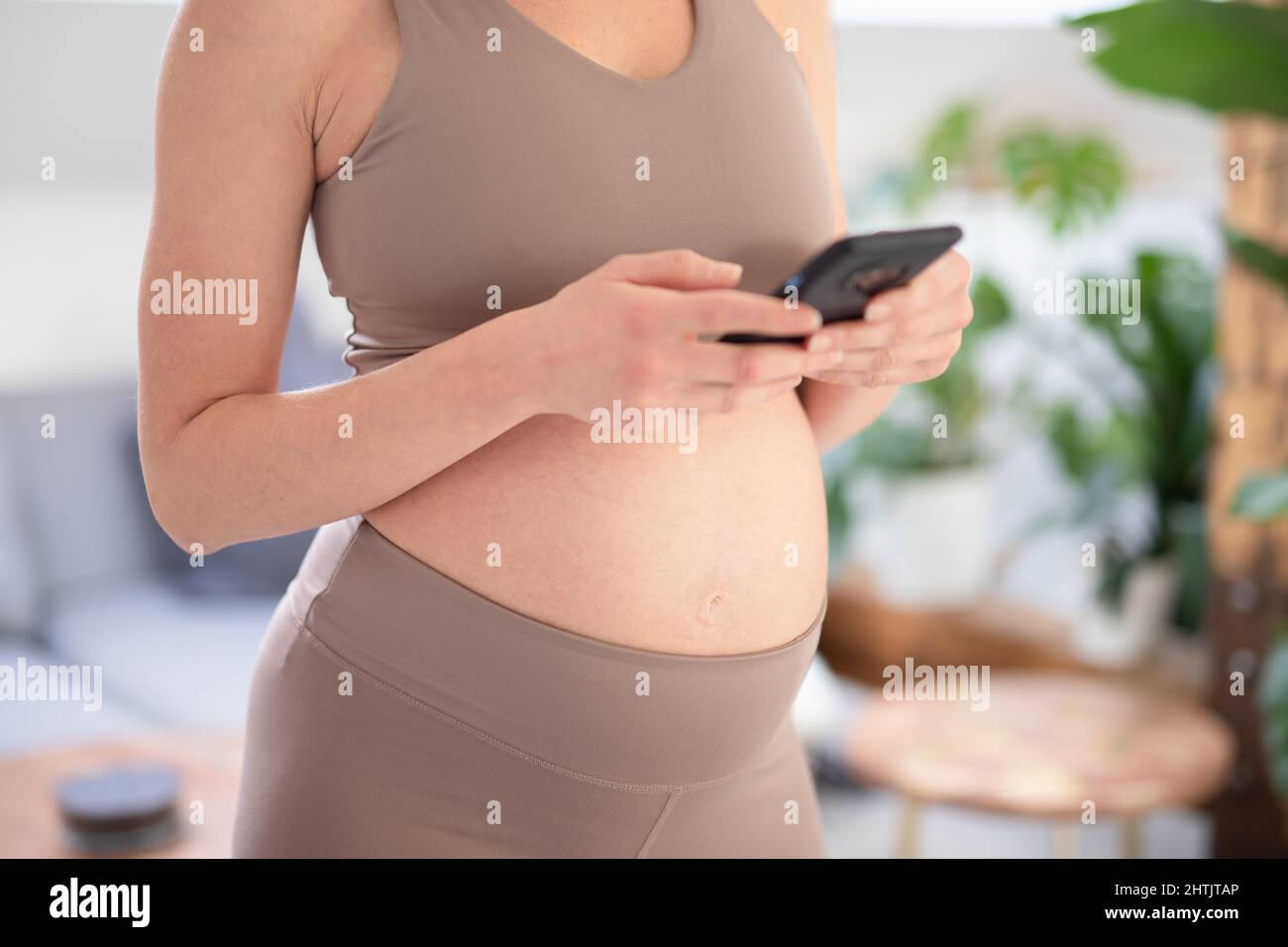 Foto ravvicinata della ventre femminile incinta. Donna che tiene e utilizza l'applicazione per smartphone mobile negli interni di casa. Gravidanza, tecnologia, online Foto Stock